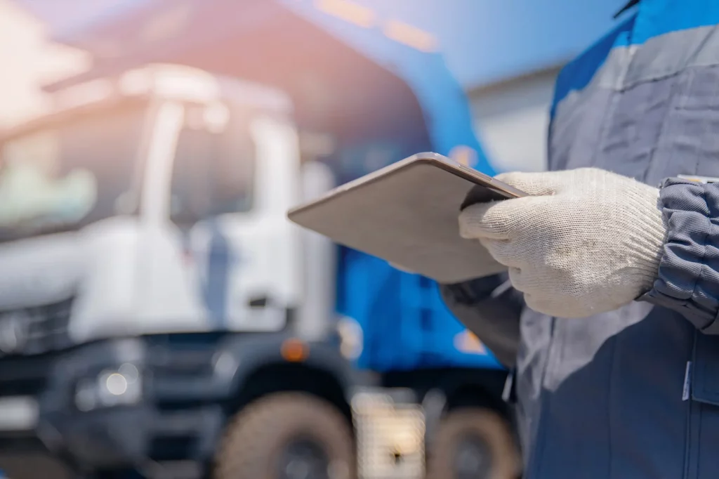 Ein Arbeiter in blauer Schutzkleidung und Arbeitshandschuhen hält ein Tablet in den Händen. Im Hintergrund ist unscharf ein großer Lastwagen zu erkennen, der auf einem Betriebsgelände geparkt ist. Die Szene deutet auf die Nutzung moderner Technologie zur Überwachung oder Verwaltung von Aufgaben im industriellen oder logistischen Bereich hin. Die Kombination aus traditioneller Arbeitskleidung und einem Tablet unterstreicht den Einsatz digitaler Werkzeuge in praktischen Arbeitsumgebungen.