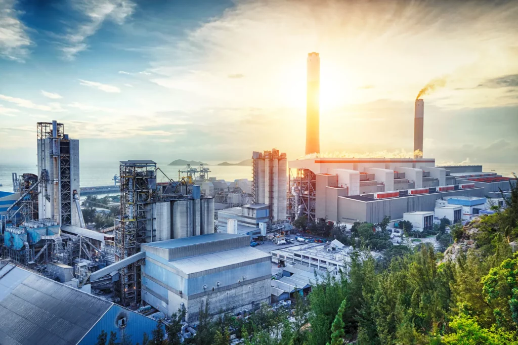 Industrieanlage bei Sonnenaufgang mit Blick auf das Meer im Hintergrund. Die Anlage besteht aus mehreren großen Gebäuden und hohen Schornsteinen, aus denen Rauch aufsteigt. Die Strukturen sind aus Beton und Metall, umgeben von zahlreichen Rohrleitungen und Maschinen. Der Himmel ist teils bewölkt, während die aufgehende Sonne die Szene in ein warmes Licht taucht. Grünflächen und Bäume umgeben das Gelände und sorgen für einen Kontrast zur industriellen Umgebung. Im Hintergrund sind kleine Inseln und Hafenanlagen zu erkennen.