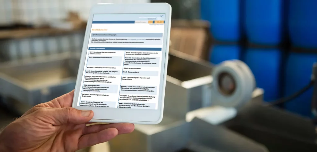 Ein Hand hält ein Tablet, das eine Anwendung mit einer Liste von Gesetzen und Vorschriften anzeigt, bekannt als das Rechtskataster des "Genehmigungsmanagers". Im Hintergrund sieht man industrielle Einrichtungen mit großen blauen Fässern. Die App auf dem Tablet zeigt eine organisierte Liste verschiedener Gesetze, geordnet nach Kategorien, um den Zugriff auf relevante rechtliche Informationen zu erleichtern. Diese Funktion unterstützt Nutzer dabei, relevante rechtliche Bestimmungen schnell zu identifizieren und einzusehen.